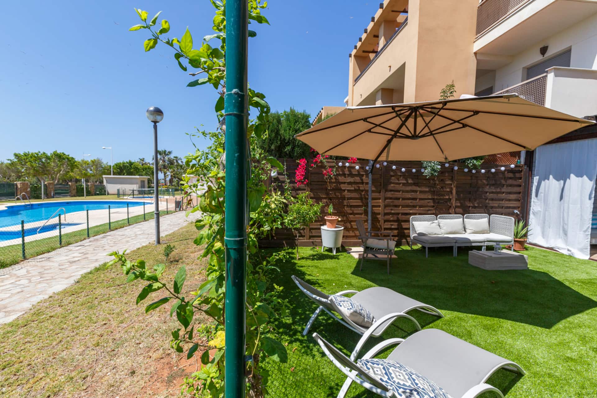 Terraza con tumbonas y sombrilla junto al jardín, con la piscina comunitaria al fondo.